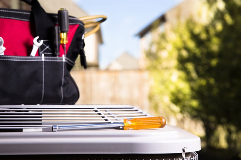 Bag of repairman's work tools, screwdriver on top of air conditioner unit outside brick home.  Service industry, working class.