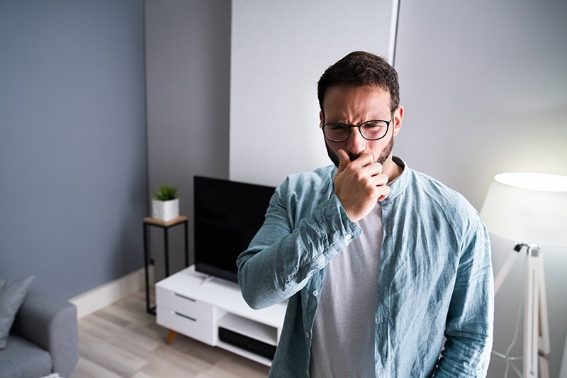 A man in glasses holding his hand up to cover his mouth or nose with an uncomfortable expression in a living room.